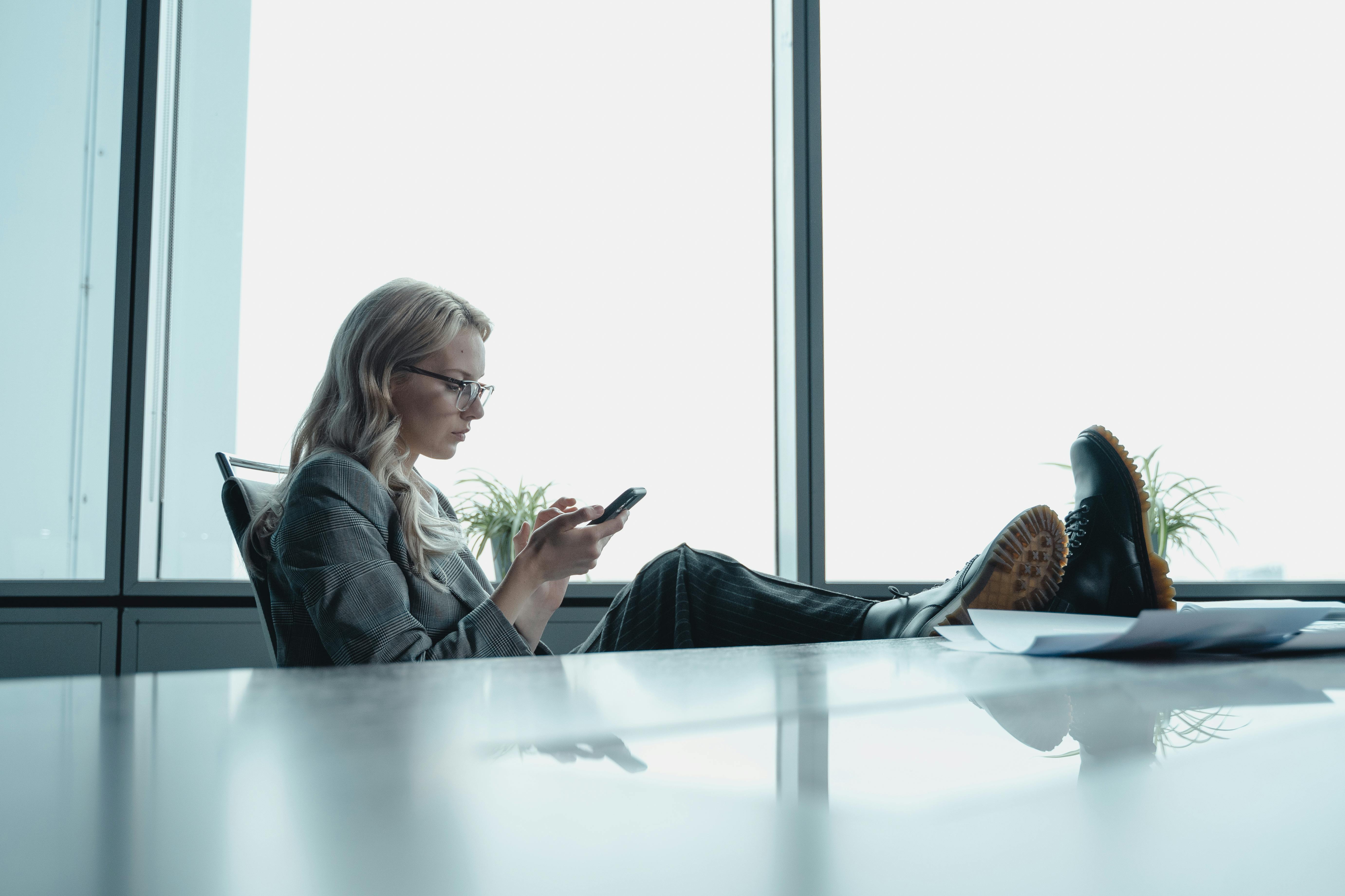 A woman in a blazer uses her smartphone, relaxing with feet up in a modern office.