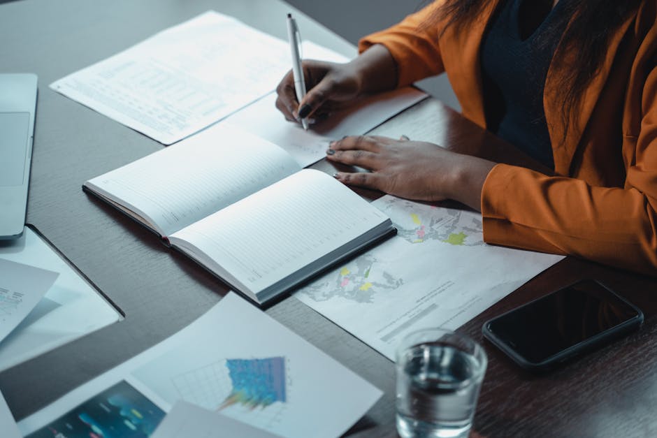 Photo by Tima Miroshnichenko A businesswoman writes in a notebook while reviewing charts and graphs in an office setting.