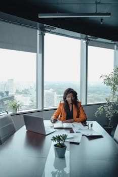 Businesswoman sitting in a high-rise office, working on a laptop with a city view.