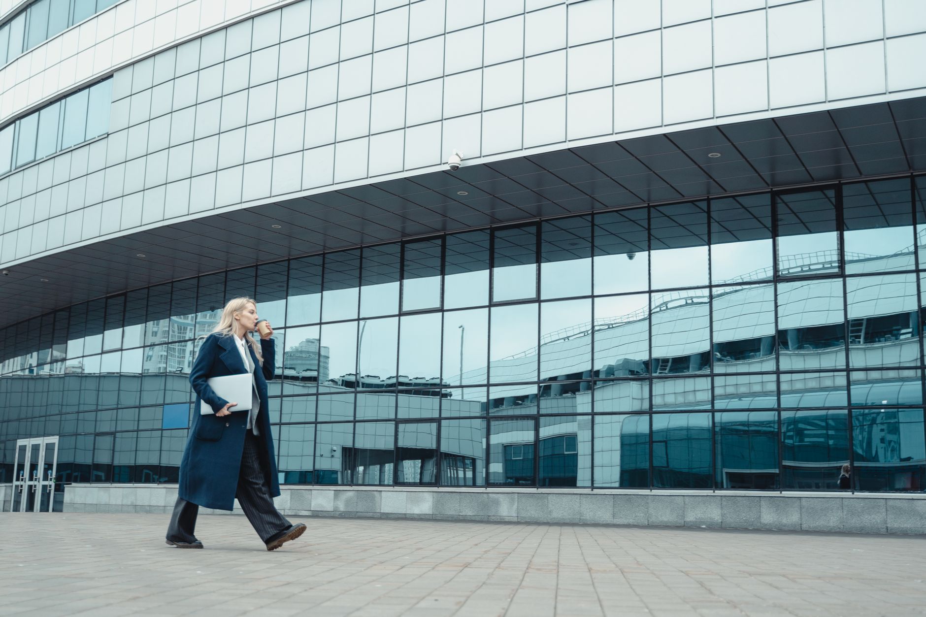 Confident businesswoman walking with coffee and tablet by modern glass-panel building, embodying urban professionalism.