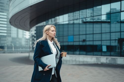 Free Confident businesswoman walking outside modern office building holding laptop and coffee. Stock Photo