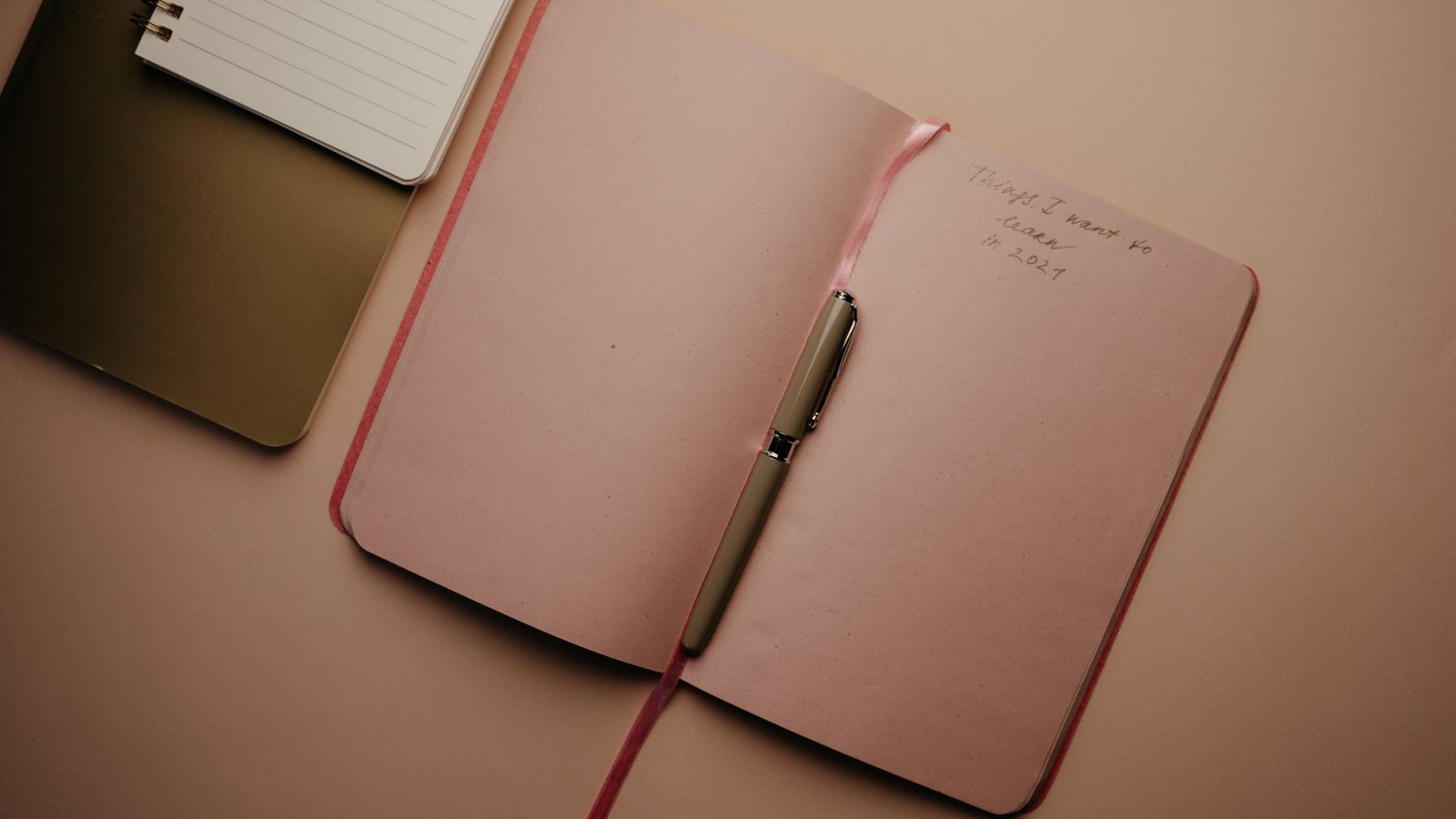 A pen resting in an open pink notebook beside a smaller lined notebook