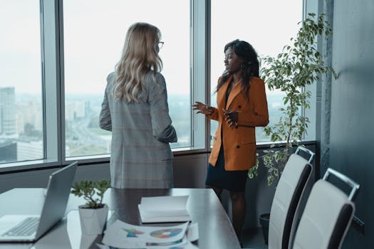 Two women discussing work in a high-rise office, showcasing diversity and teamwork.