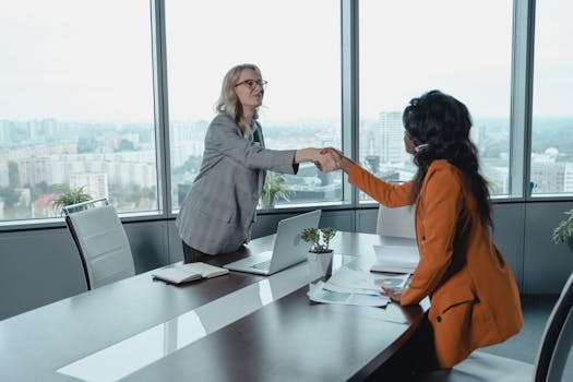 Two businesswomen shaking hands during a meeting in a high-rise office with a cityscape view.