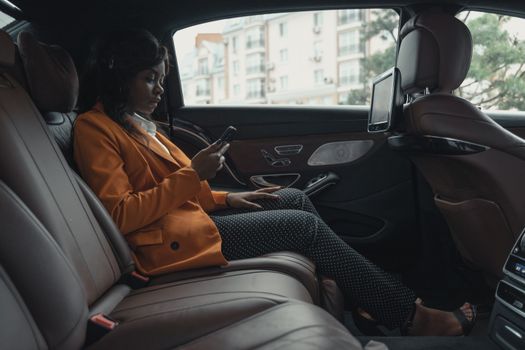Professional woman in orange jacket checking phone while sitting in car's backseat.