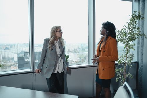Free Two businesswomen conversing in a modern office with a city view, promoting corporate diversity. Stock Photo