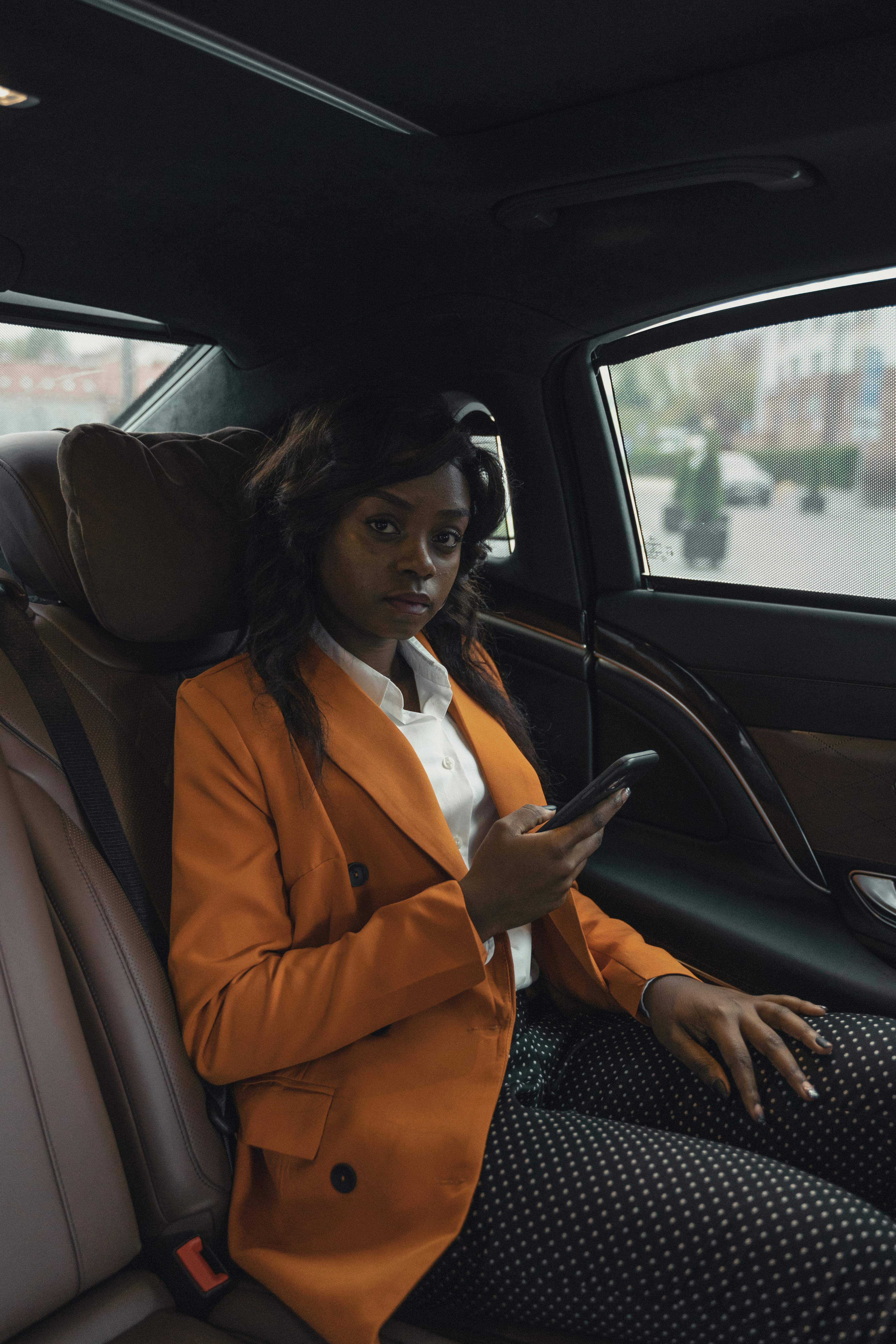 woman in orange blazer sitting on car holding her phone