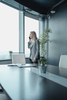 Professional woman in a stylish suit engaged in a phone call in a sleek, modern office setting with large windows.