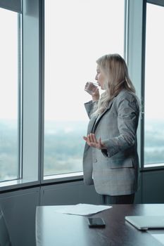 一位穿着西装的女商人站在现代办公室的窗前喝水