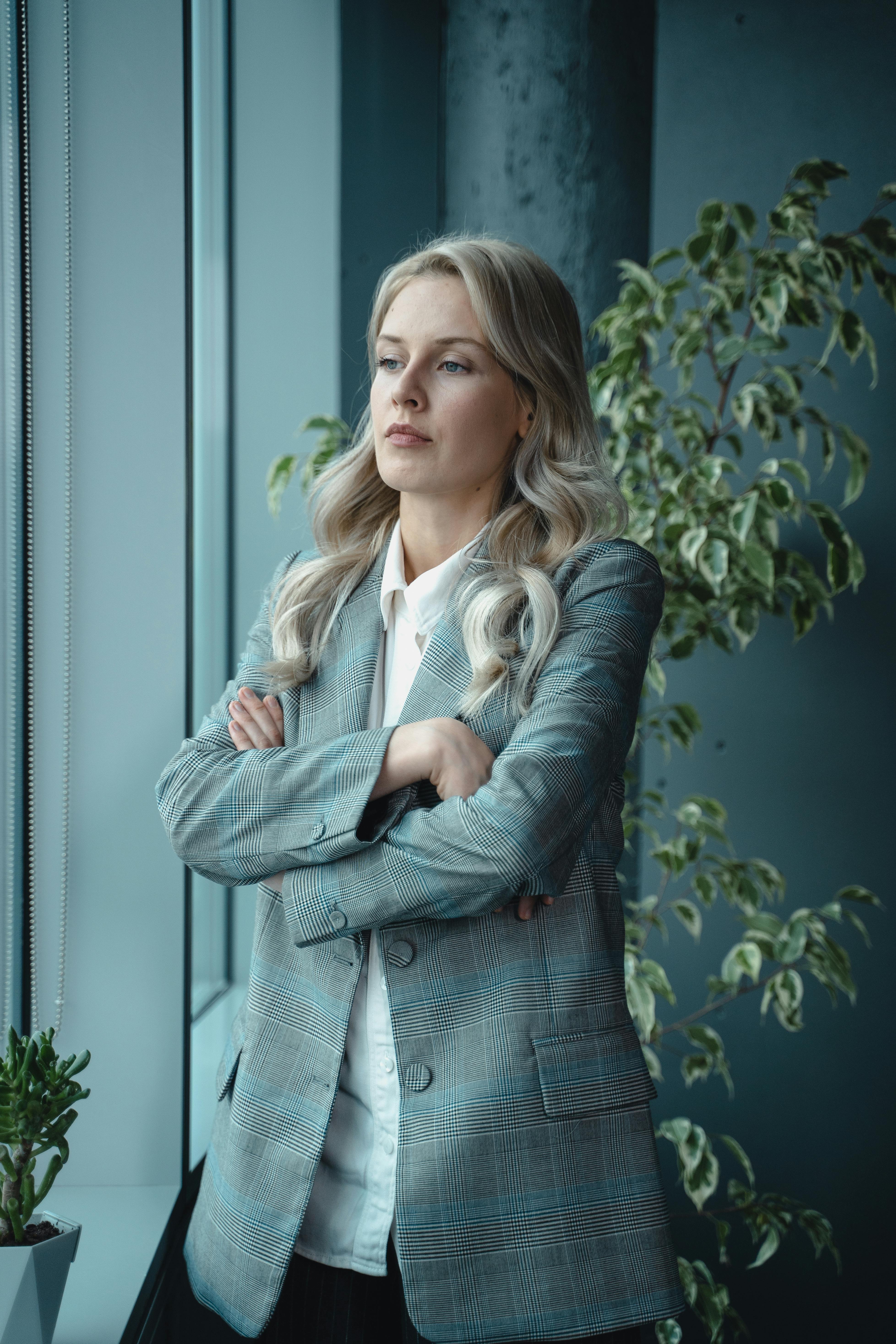 woman in gray blazer standing near green plant with a serious look