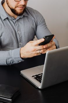 一名成年男子在办公桌前用笔记本电脑工作时，同时使用着智能手机