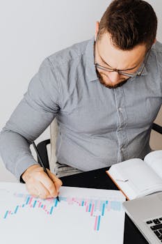 Focused businessman in gray shirt analyzing a financial chart at his office desk, emphasizing data-driven decision making.