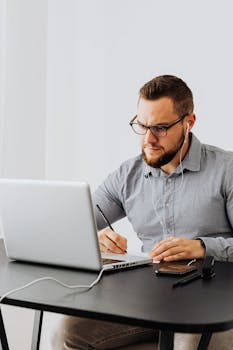 Focused man with a beard uses laptop and writes notes, working from home.