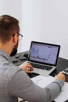 Man working on a laptop analyzing financial data with graphs in an office environment.