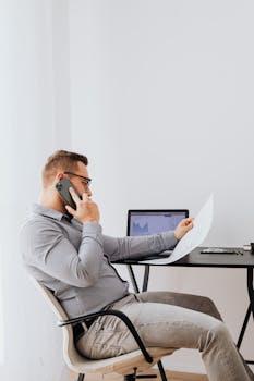 Business professional seated at desk, reviewing documents while conversing on phone.