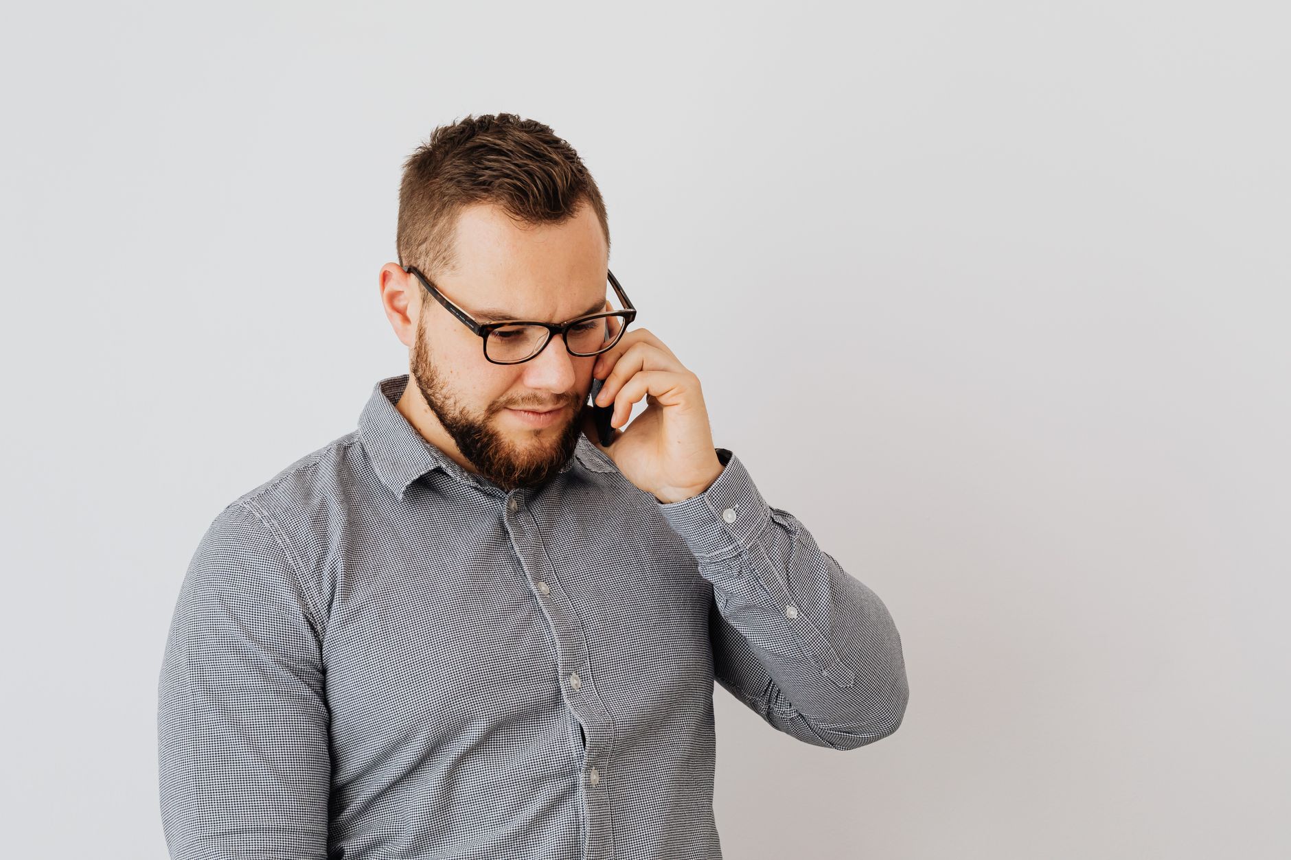 Young man with beard in glasses and gray shirt talking on smartphone indoors.