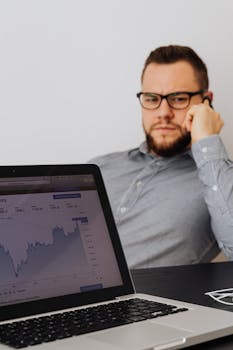 Man in office evaluating stock market graphs on his laptop while on the phone.
