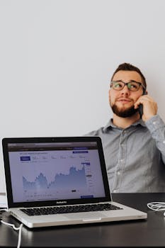 A man talks on the phone while viewing cryptocurrency graphs on a laptop in an office setting.