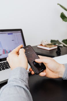Person analyzing stock market data on smartphone, with laptop in background.