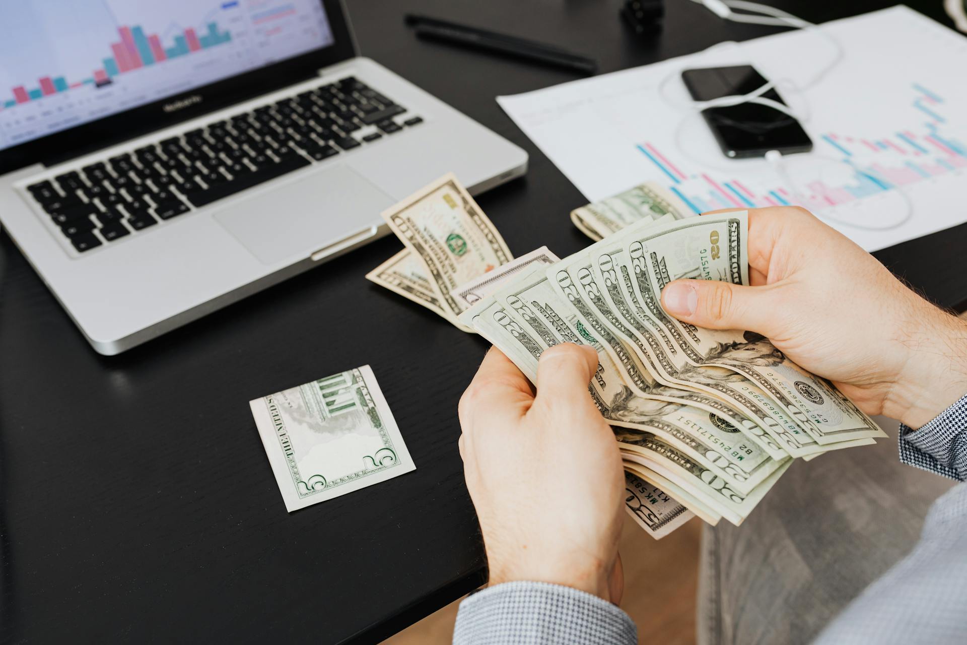 Person counting dollar bills at desk with laptop and financial charts, symbolizing finance management.