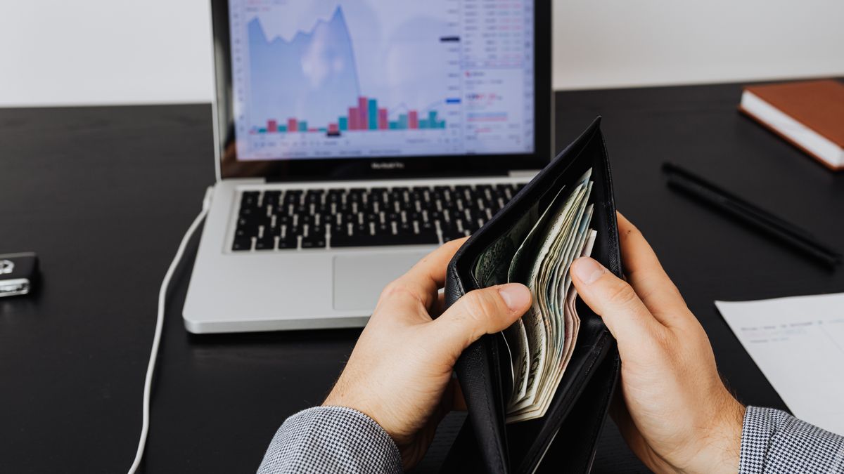 Man holding a leather wallet with bills in front of a laptop displaying financial graphs.