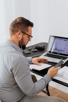 Business professional analyzing financial graphs on laptop while using smartphone in a home office.