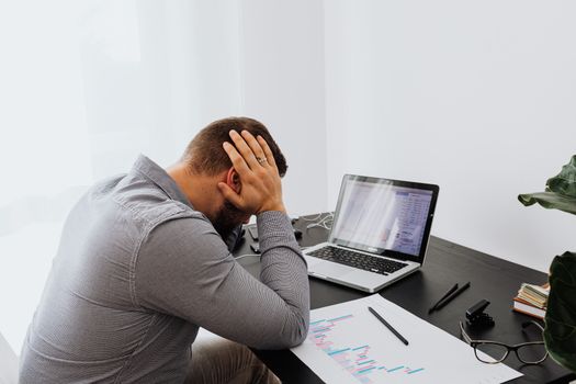 一个男人双手抱头，看着办公桌上的笔记本电脑和图表