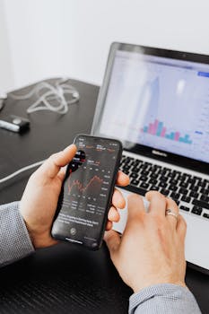 Hands holding smartphone analyzing stock market data next to laptop displaying charts.