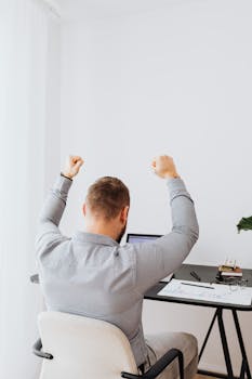 A businessman celebrates success raising arms while sitting at his desk.