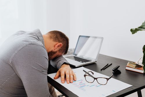 Free A man in a gray shirt sits tiredly at a desk with a laptop and documents, showing signs of workplace stress. Stock Photo