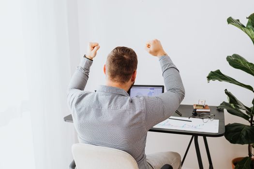 Back view of a man cheering with arms raised while sitting at a desk.