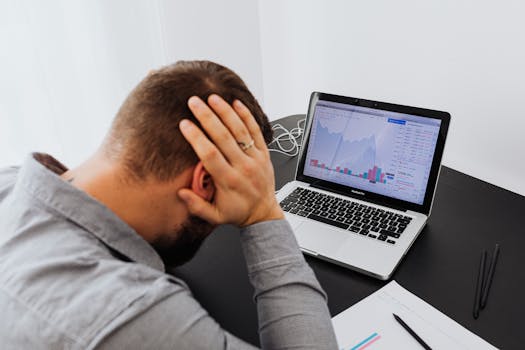 Stressed man at desk looking at declining stock charts on laptop, indicating financial loss.