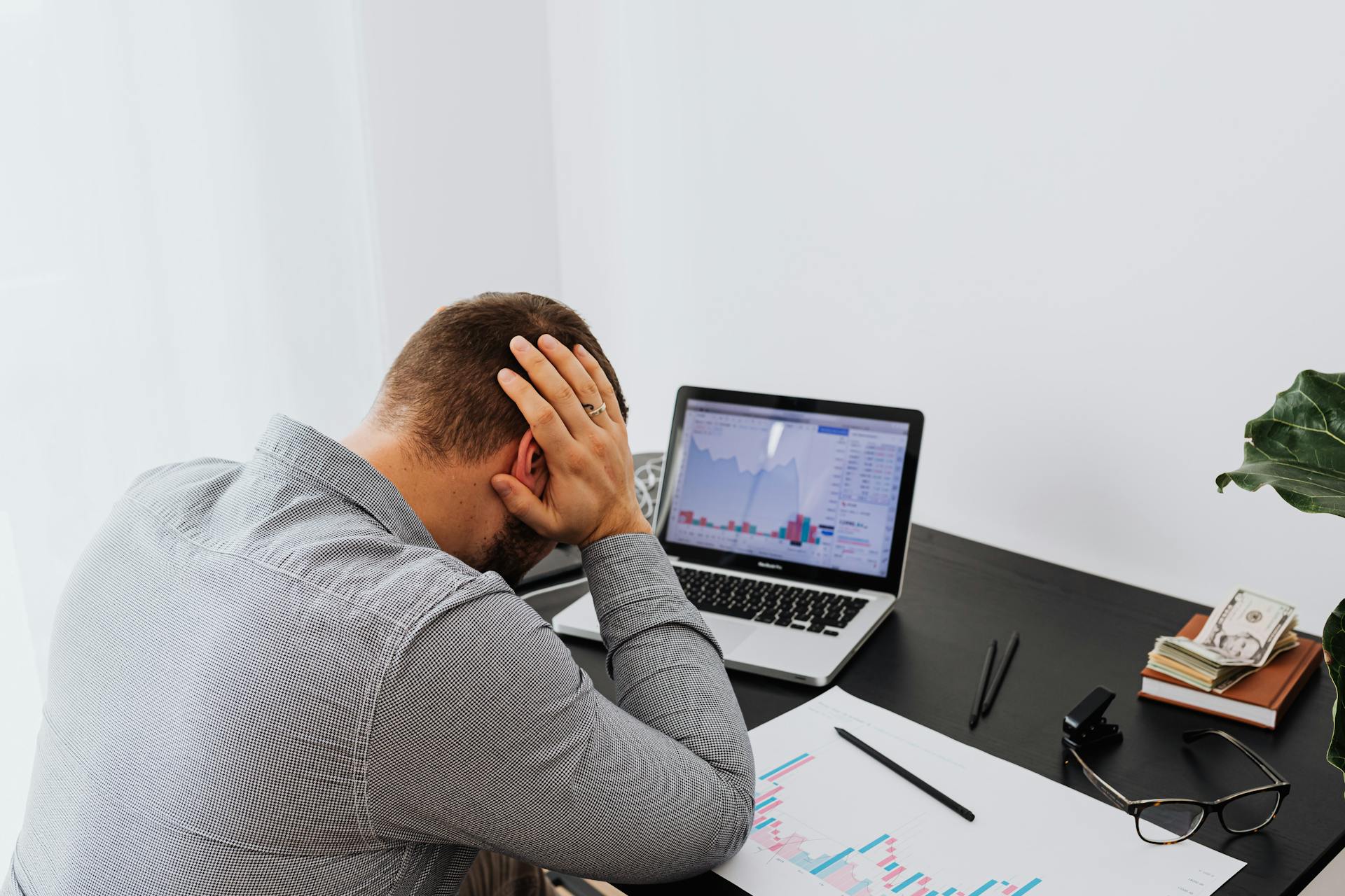 A businessman holds his head in frustration while sitting at a desk with a laptop and financial charts.