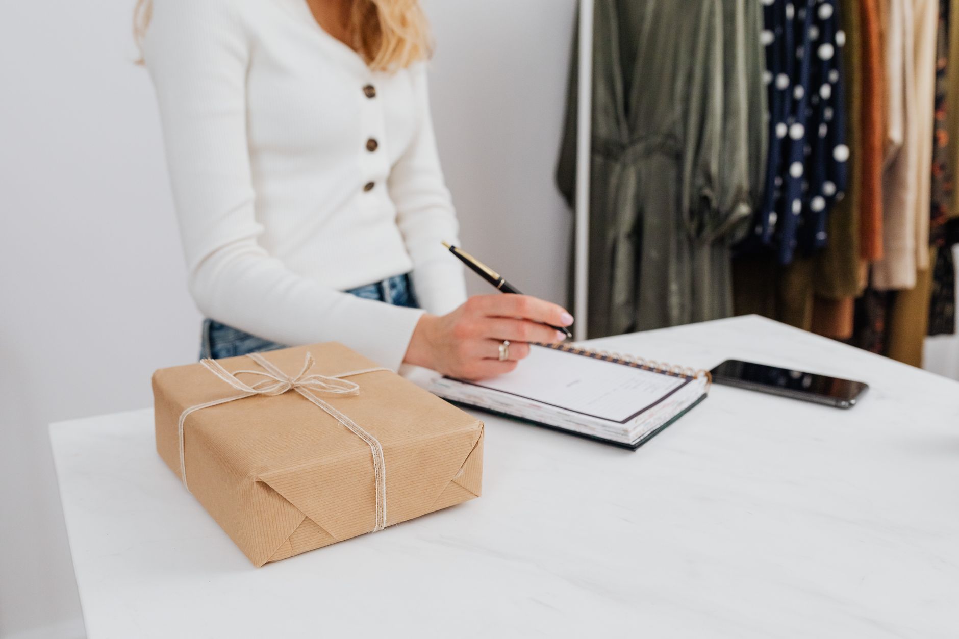 Woman writing in notebook beside a wrapped gift box on a white table indoors.