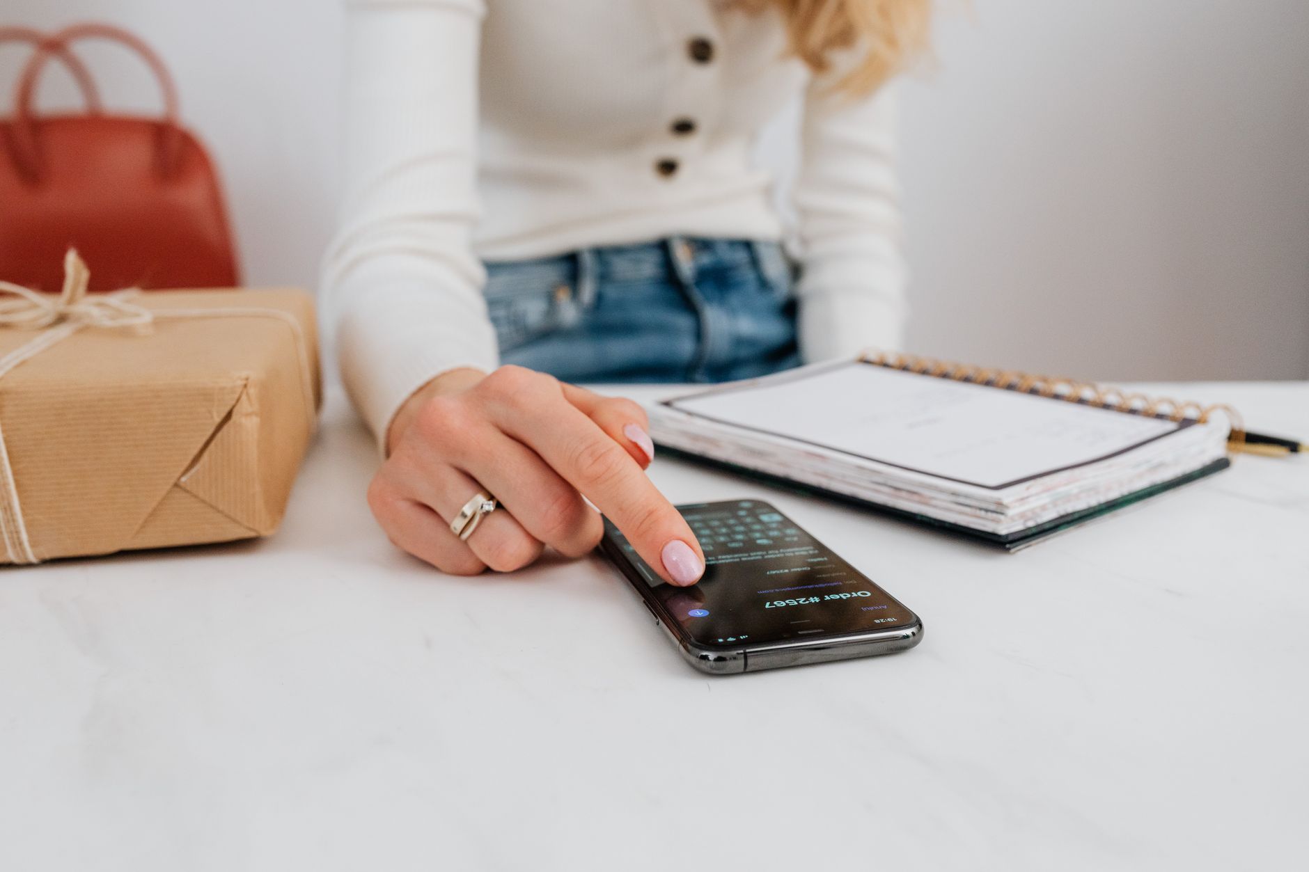 A woman interacts with her smartphone on a white table, next to a gift box and notebook.