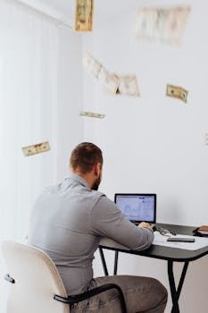 A man sits at a desk with a laptop and floating currency, symbolizing financial growth.