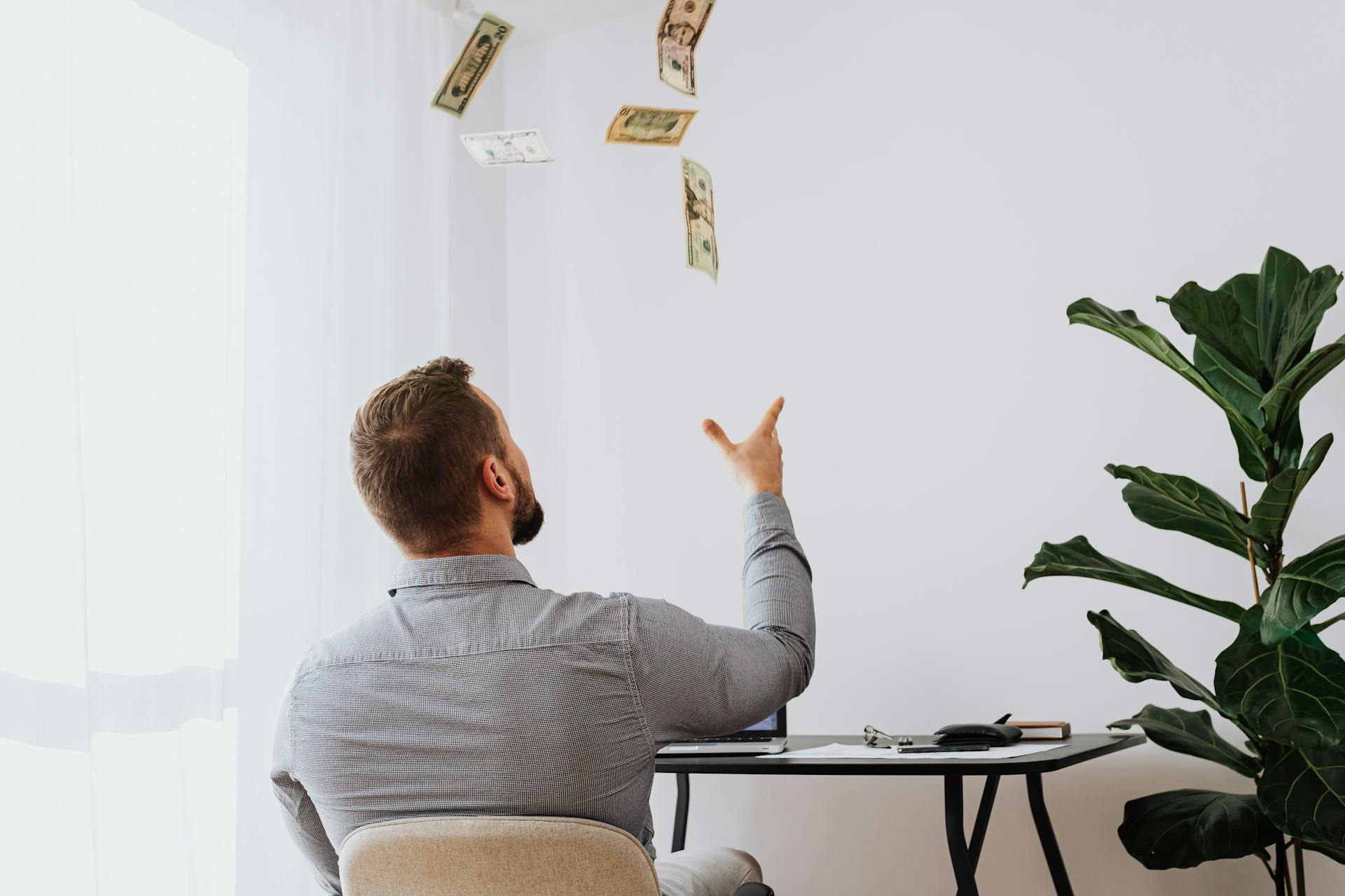 Man throwing cash in the air while sitting at a desk indoors, symbolizing financial success.