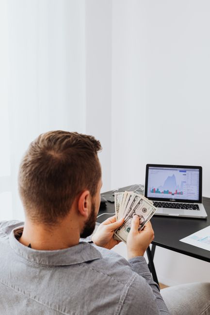 Businessman holding money while analyzing stock market data on laptop at home office setup. Businessman holding money while analyzing stock market data on laptop at home office setup.