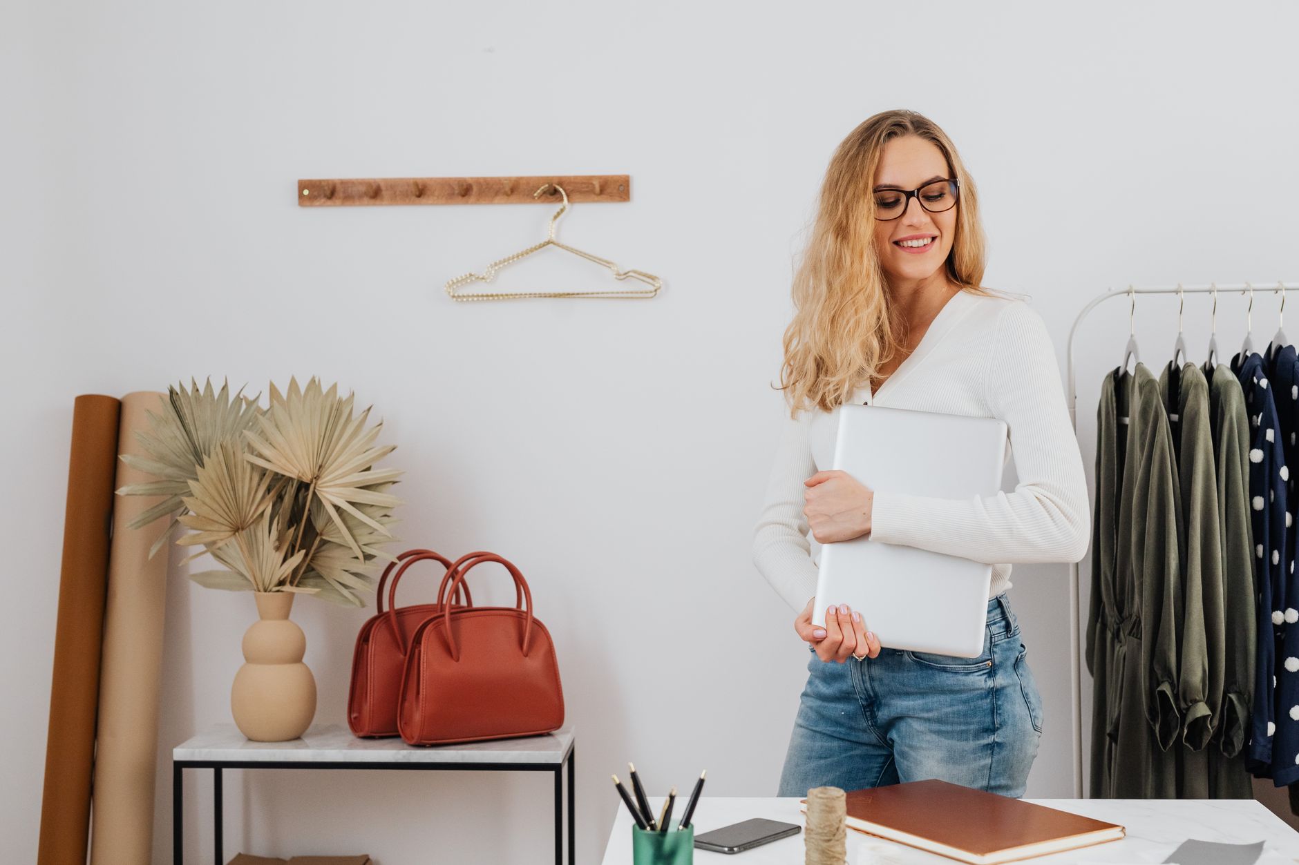 A fashionable woman holding a laptop stands in a chic boutique setting with clothes and accessories.