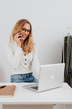 Blonde woman using laptop and phone for remote work at home office.