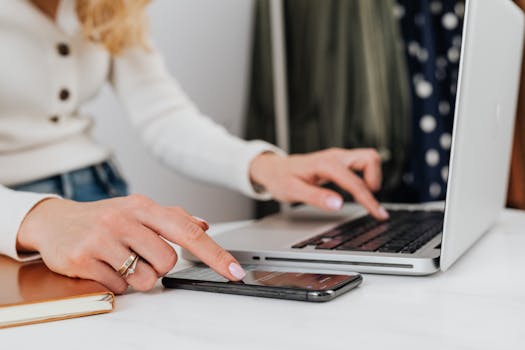 Close-up of a woman's hands using a laptop and smartphone simultaneously on a desk.