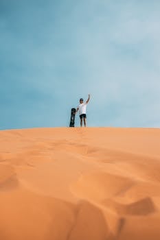 A lone adventurer poses at the top of a sand dune in Utah, ready for sandboarding under a clear blue sky.