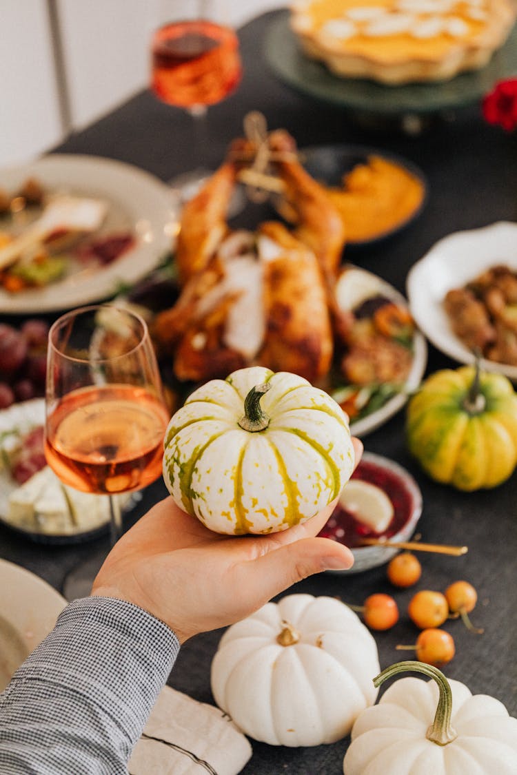 Person Holding A Small Ornamental Pumpkin Over A Table With Food 