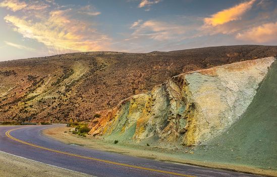 Curved road through colorful rock formations and hills under a vibrant sky.