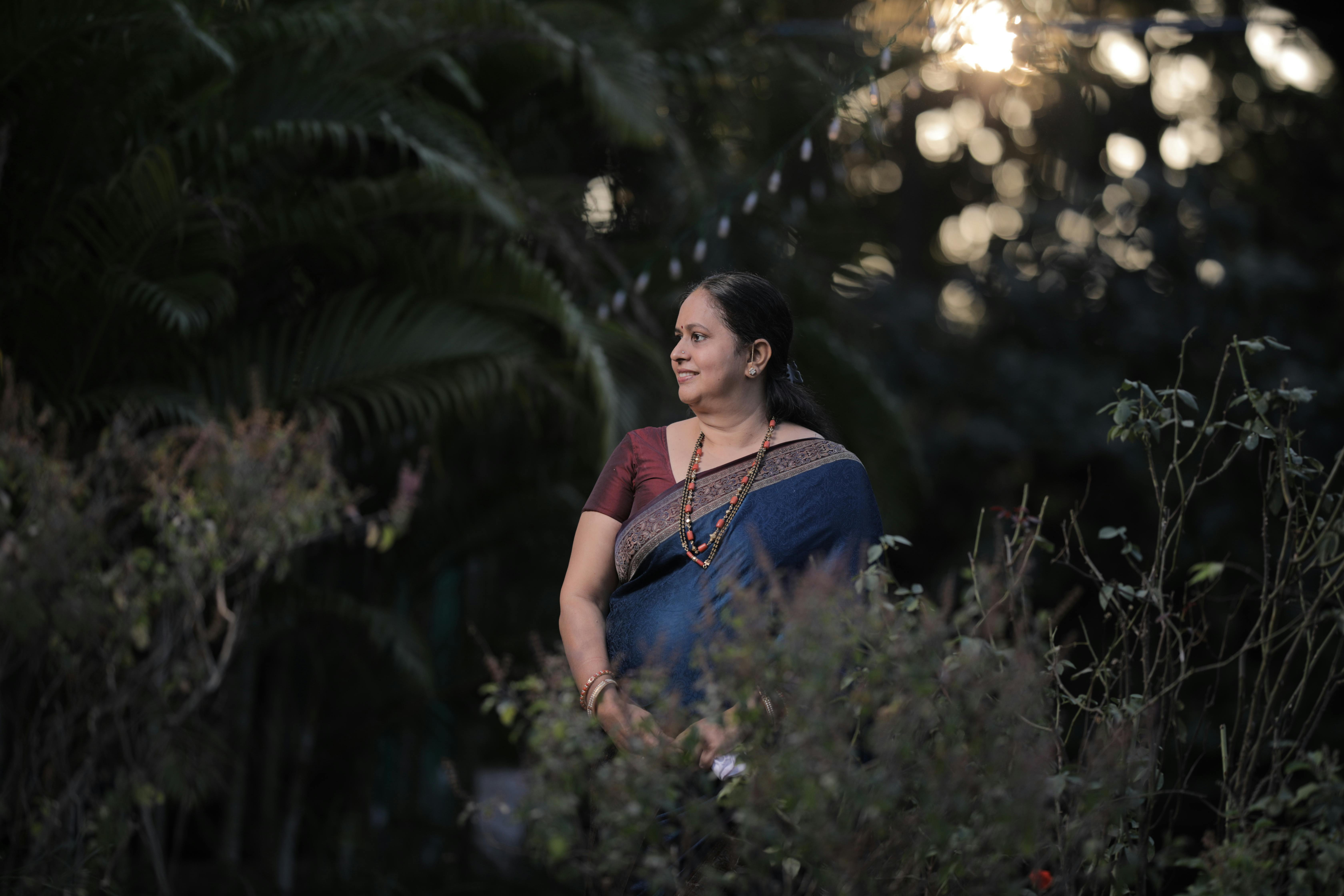 A smiling woman in a saree looks away while standing in a park.