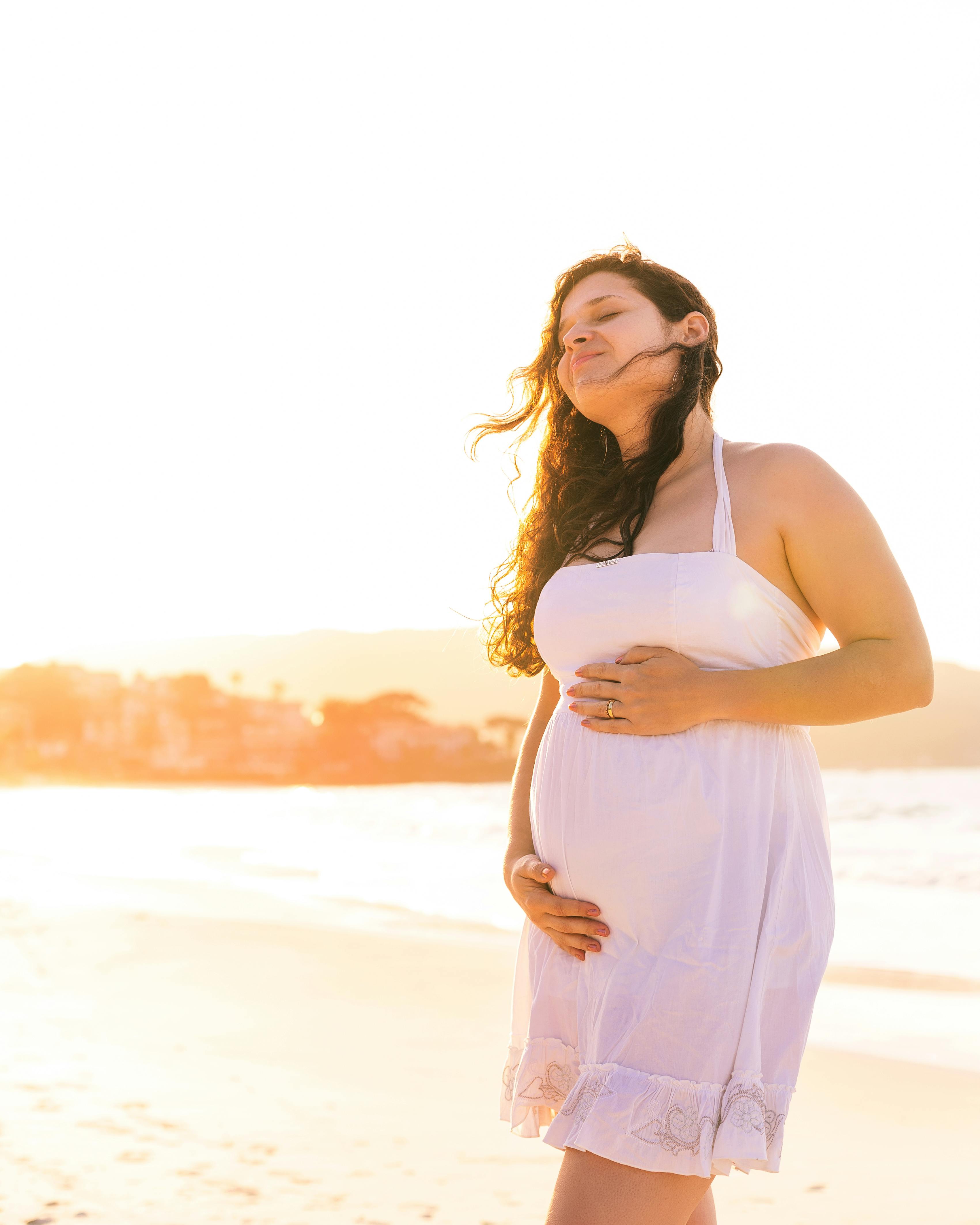 pregnant woman in white spaghetti strap dress standing on beach