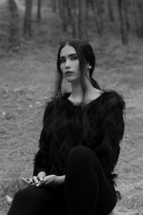 Black and white calm attractive female wearing fluffy dark sweater sitting on ground in woodland and looking at camera