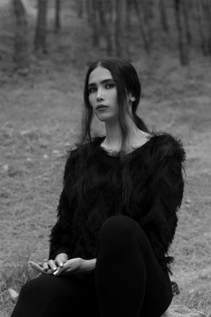 Black and white calm attractive female wearing fluffy dark sweater sitting on ground in woodland and looking at camera