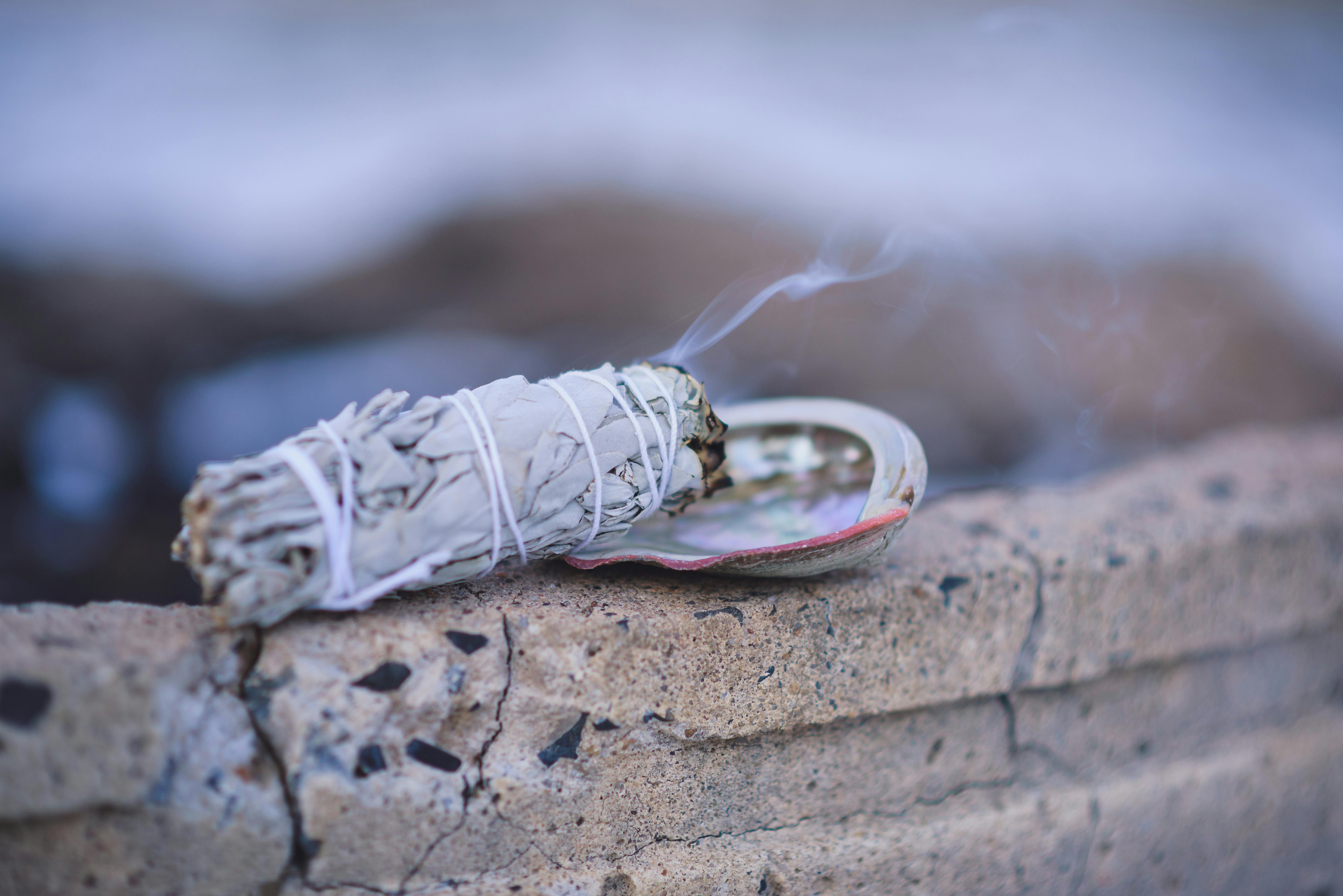 A close-up view of burning sage in a smudging ritual, releasing smoke over a stone surface.