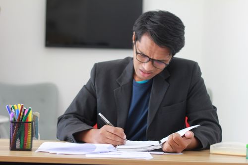 Free Adult male in a black suit takes notes at a cluttered office desk, looking stressed. Stock Photo